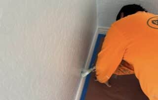 A professional painter carefully applies white paint to the baseboard trim of a textured wall.