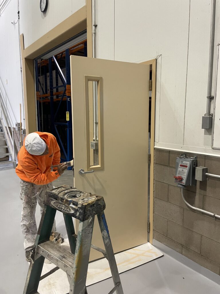 Commercial Interior Door Painting Services A painter applies a fresh coat of beige paint to a commercial building's interior door.